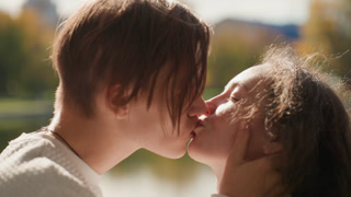 Caucasian couple kissing by lakeside sunlight, aspiring artists share playful embrace with hands cradling face, soft autumn backdrop