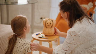 Mother and white girl crafting pumpkin at small table, playful smiles, woman guiding brush and marker, cozy livingroom scene, warm daylight