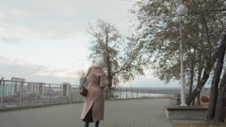 Caucasian woman walking away from camera along waterfront, coat and beanie, autumn leaves falling, distant skyline and harbor, slow