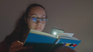 Young woman reading by phone light, latenight student studying notebook in dim bedroom, wearing glasses and curly hair, colorful dotted