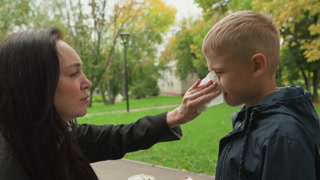 White boy mother firm nose wipe before school, park setting with trees, stern but caring expression, mother pointing finger and giving
