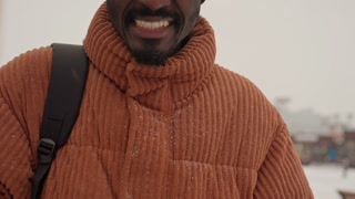 Black man smiling in snowy street wearing orange chunky sweater black beanie glasses and mittens with backpack gentle snowfall downtown