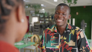 Black man and woman on date sharing shy smiles in bright modern cafe, young student man offering compliment with attentive gaze, colorful