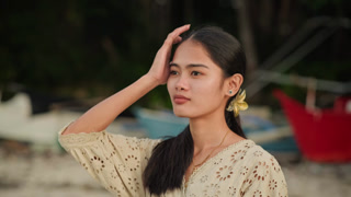 Asian woman at beach closeup, side profile with tropical flower in hair, subtle necklace glint, adjusting hair and gazing toward horizon