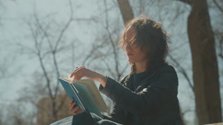 Woman reads blue book with colorful circles on cover outdoors surrounded by bare trees and sunlight. Woman holds open pages in daylight