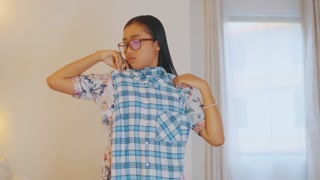 Confused young woman trying on plaid shirt, making decision on outfit at home.