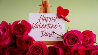 Happy Valentine's Day greeting card displayed on wooden easel surrounded by vibrant pink roses and decorative red heart shapes on soft
