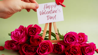 Happy Valentine's Day greeting card displayed on wooden easel surrounded by vibrant pink roses and decorative red heart shapes on soft