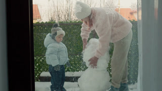 A mother helps her young boy make a snowman in their apartment yard. They shape the snow and have fun together on a cold winter day while laughing.
