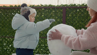 A mother and her son enjoy winter by making a snowman in the yard. They gather snow and shape it together while having fun in their apartment building's area.