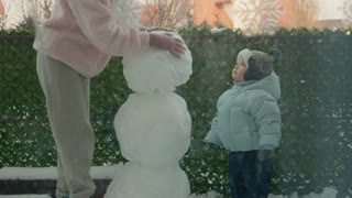 A mother and her little boy are outside in the snow making a snowman. They work together in the yard of their apartment building on a winter day.