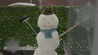 A snowman is built in the yard of an apartment building. It holds a broom and wears a hat. Snow covers the ground in a typical winter scene.