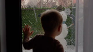 A boy stands by the window and watches a snowman in the yard. The snowman has a hat and scarf and looks fun. Snow is all around, creating a winter scene.