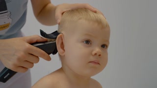 A mother skillfully cuts her young son's hair at home using clippers and scissors while he sits calmly. The atmosphere is relaxed and nurturing during the haircutting process.