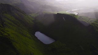 Aerial view of a serene lake in a green valley, a winding river reflecting sunlight, and rugged volcanic terrain under a soft sunset glow in Iceland.