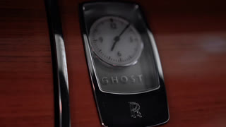 Close up pans across a chrome trimmed analog clock in Rolls Royce Ghost wood veneer. GHOST wordmark and double R emblem appear, with warm tones and shallow depth.