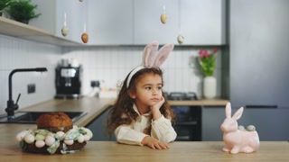 Sad easter little girl in bunny ears sitting in the decorated kitchen alone. Disappointed child. Kid feeling unhappy on spring holiday
