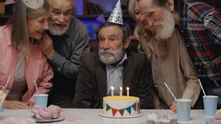 Happy Funny Senior Man in Birthday Hat Blowing Out Candle on Birthday Cake, Enjoying Celebrating Together with Other Elderly People at Cozy