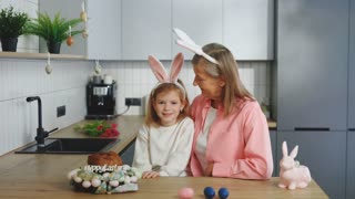 Happy Easter. Grandmother and Granddaughter spending time together, wearing Bunny ears, Looking at Camera in Kitchen. Portrait of the
