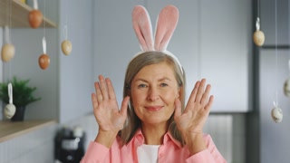 Portrait of smiling senior lady wearing bunny ears headband playing peek a boo game standing at the kitchen at home. Happy elderly woman