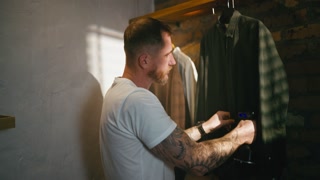 Close-up of a concentrated man in a white T-shirt and with tattoos steams his shirt using a steamer while preparing to go out at work on a