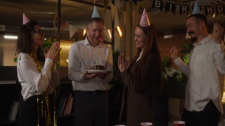Happy girl in business clothes and with a festive cap blows out candles on a birthday cake during a corporate birthday party in the office
