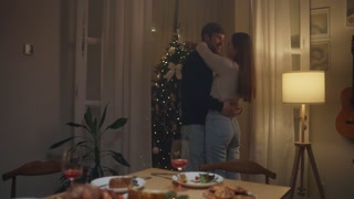 Couple shares a romantic moment by the Christmas tree during a cozy dinner in their warmly lit home setting