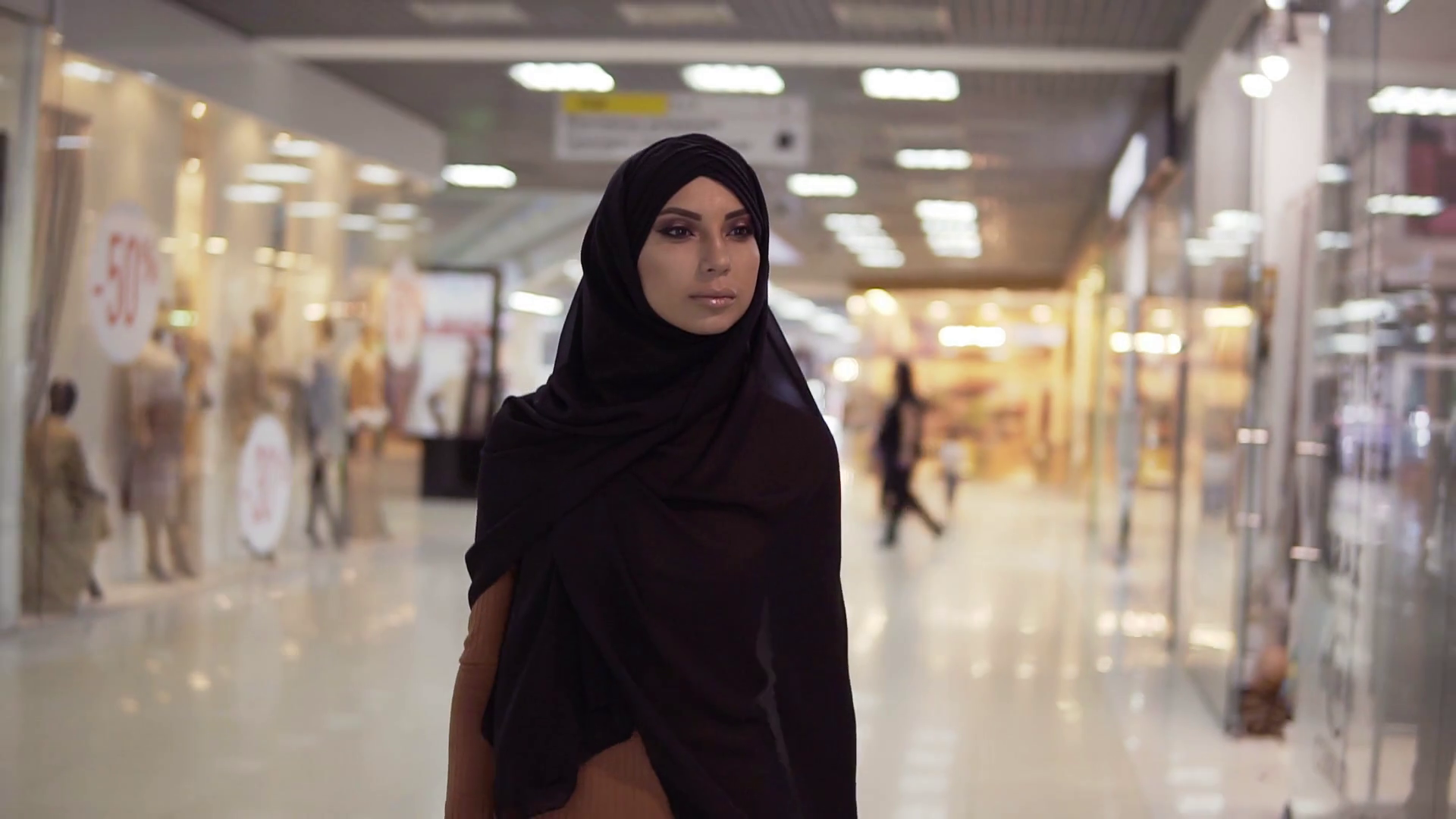 Beautiful muslim woman in hijab walking by big mall and looking for sales through windows. Atractive islamic girl doing window shopping