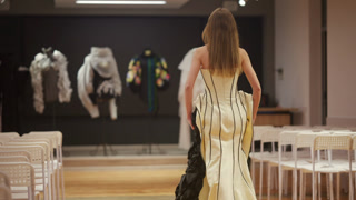 Back view of a female model walking in a structured corset style dress with vertical black boning lines, presented in a minimalist indoor fashion runway