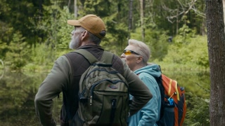 Back view shot of happy senior Caucasian couple with camping backpacks enjoying scenery stopping by lake during their hike on chilly summer day