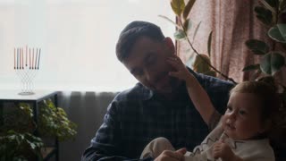 Tilt down shot of loving young Jewish father playing with his joyful toddler son sitting on chair in modern apartment with hanukkiah decorated with colored candles in background