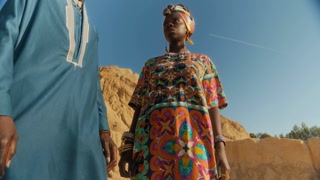 Low angle view of young Black man and woman wearing modern ethnic clothes standing in desert, looking at camera