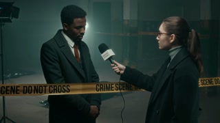 Medium lowkey shot of young Caucasian female reporter asking young Black male detective agent questions about murder case standing at crime scene isolated by yellow police tape