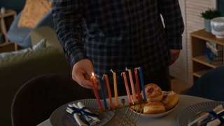 Tilt down shot of young Jewish man holding burning shammash and lighting up candles on hanukkiah while singing holiday hymn Maoz Tzur, celebrating Hanukkah at home