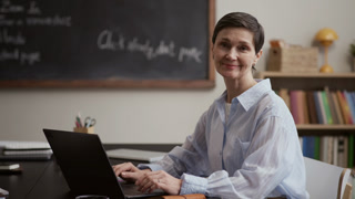 Waist up portrait of mature Caucasian female student smiling at camera as sitting at desk against chalkboard and using laptop while studying in modern classroom learning environment