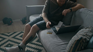 Tilt shot of Caucasian man wearing ankle monitor sitting on couch with laptop and sorting bills at home while feeling overwhelmed by financial responsibilities