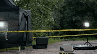 No people shot of outdoor murder scene with dead body under plastic bag on ground and professional lighting equipment set up around it in park at night