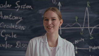 Chest up portrait of young Caucasian female medical student smiling at camera while standing against chalkboard with written theory and graph wearing white lab coat