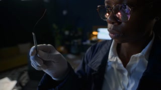 Young African American female police officer examining hair, collecting trace evidence using tweezers into transparent zip bag, working at crime scene in office