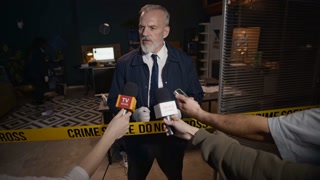 Medium long shot of mature Caucasian male police officer being interviewed by TV reporters at crime scene isolated with yellow barricade tape