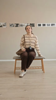 Vertical full POV shot of young Caucasian woman wearing audio guide earphones taking seat on bench in modern art gallery and looking at camera