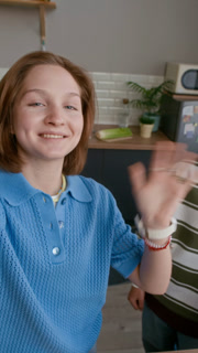 Vertical handheld POV shot of two Caucasian teenage bloggers filming funny cooking show at home, girl holding camera while boy smiling with orange peel in his mouth in bright kitchen