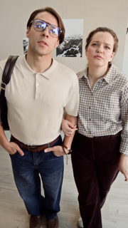 Vertical POV medium long shot of young Caucasian couple discussing artworks while looking at camera, standing in exhibition hall in modern art gallery