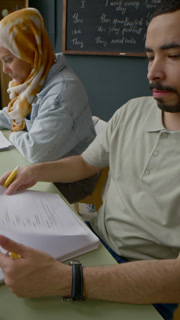 Vertical of group of diverse students taking papers from Caucasian female teacher while seated at desk with study materials in modern classroom
