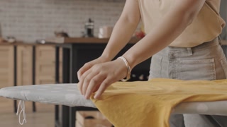 Tilt up slowmo shot of young woman ironing T-shirt on ironing board at modern apartment