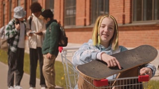 Medium slowmo portrait of cheerful Caucasian gen z girl with skateboard waving at camera sitting in shopping cart outdoors in summer while her diverse friends chatting in blurred background