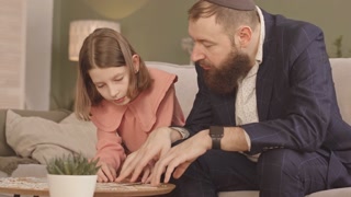 Jewish father wearing kippah and his elementary school girl solving jigsaw puzzle together while sitting on sofa at coffee table in living room