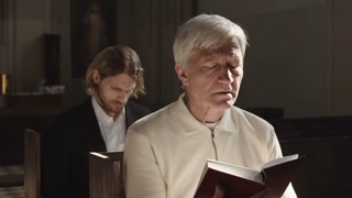 Medium shot of two Caucasian men of different age sitting on wooden benches in Lutheran church and reading Holy Bible during Sunday service