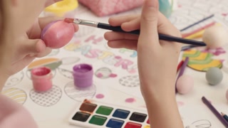 Slow-motion close-up of child coloring Easter egg in pink with brush at table with paints, coloring pages, and Easter decorations