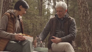 Slow motion of mature couple conversing over lunch in serene forest. Man pouring tea into thermos mugs.